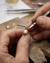 Close-up of hands working on a gold ring with a diamond using tools on a wooden surface.